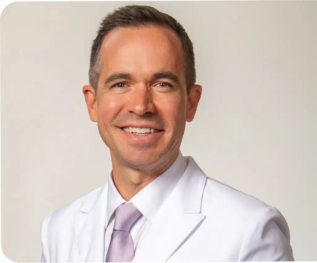 Professional headshot of male dentist in white coat and purple tie against neutral background.