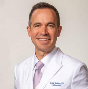 Professional headshot of male dentist in white coat and purple tie, smiling at camera.