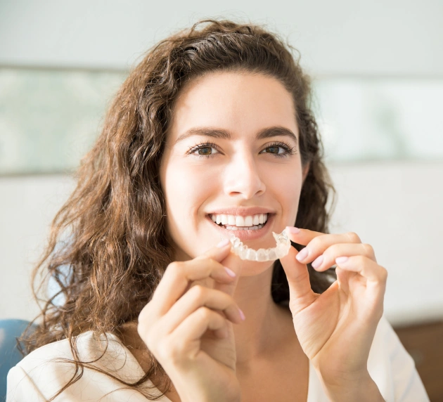 Woman smiling while holding clear dental aligners in modern dental office setting.