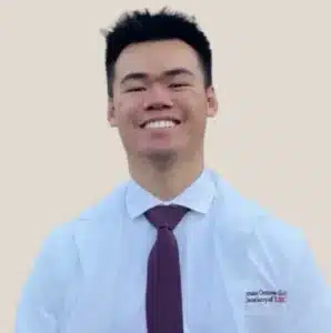 Professional headshot of smiling male dental care provider in light blue shirt and burgundy tie against neutral background.